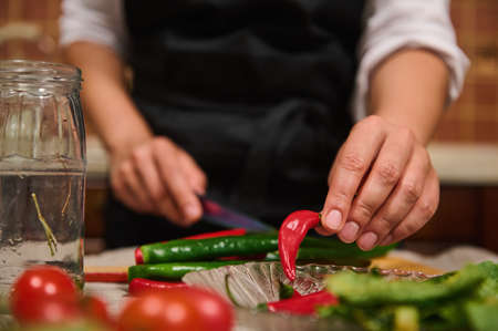 Details: Chefs hand takes red chili peppers out of a plate while slicing raw spicy ingredients during canning food. Raw fresh vegetables are on the table on the blurred foreground. Culinary. Kitchenの写真素材