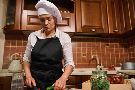 Portrait of multi-ethnic charming middle-aged woman, confident housewife in white chef cap and black apron, cooking at home kitchen, slicing raw vegetables on a chopping board. Cuisine, culinaryの写真素材