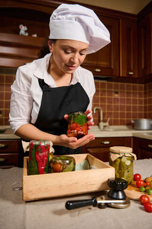 Beautiful woman, pleasant housewife in a white chefs hat and a black apron, stands at the kitchen table and puts a jar of fermented cherry tomatoes upside down on a wooden box with canned vegetablesの写真素材