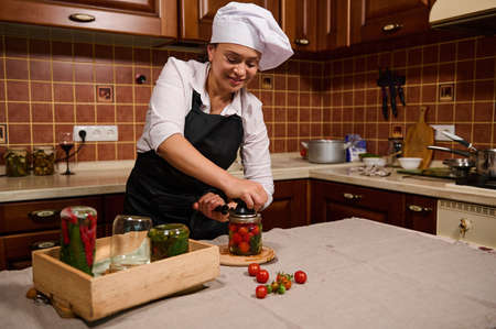 Delightful multi-ethnic young woman, housewife wearing white chefs cap and black apron, using...