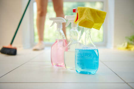 Soft focus on a yellow rag on detergents, household chemicals and cleaners standing on the floor against a blurred background of a housewife sweeping the floor in light home interior. Spring cleaningの写真素材
