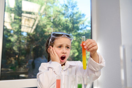 Smart Caucasian schoolgirl watching fascinating wonderful chemical reaction, between multi-colored chemicals and reagents, taking place in a test tube that she holds in her hands at a chemistry lessonの写真素材