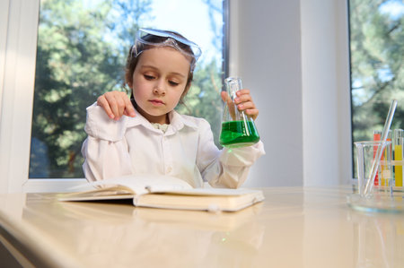 Thoughtful Caucasian little girl in safety goggles and white lab coat, concluding science experiments in chemistry class, reading text and holding a flat bottomed flask with green chemical solutionの写真素材