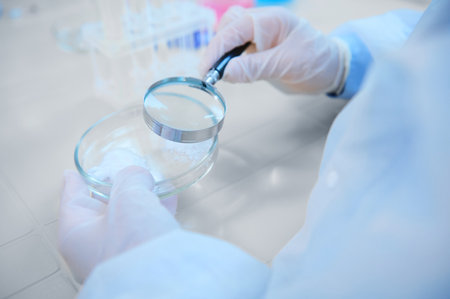 Selective focus: Loupe with magnifying glass in a scientist pharmacologist hands, examining pharmacological substance on Petri dish, sitting at table with empty test tubes on tripod in medical biolab.の写真素材
