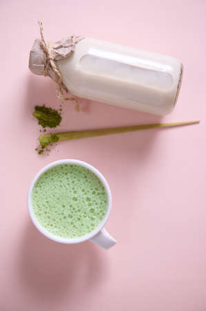 Flat lay. Vertical studio shot of a white ceramic cup of whipped matcha latte with foam, bamboo scoop with powdered green tea, bottle of healthy organic wholesome raw vegan milk over pink backgroundの写真素材