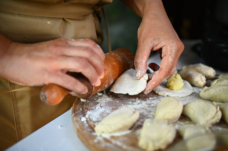 Details: Hands of a chef in beige apron, using a rolling pin, rolls out dough on a wooden board,...