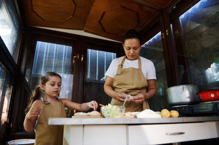 Beautiful child, a little girl in a beige chefs apron, standing next to her mother in the kitchen of...