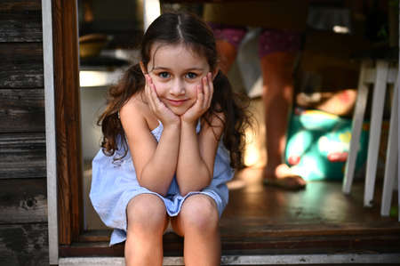 Confident portrait of an adorable 5 years old child, cute little girl, in a blue sundress, sitting on the doorstep of a country kitchen and looking at camera. Beautiful people. Children. Childhoodの写真素材