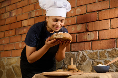 Latin American woman baker confectioner in black chefs apron and white cap, stands against brick...