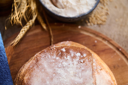 Cropped view. Still life with a loaf of wholesome whole grain homemade sourdough wheaten bread with crust, on a wooden cutting board, next to white flour in blue ceramic bowl and spikelets of wheatの写真素材