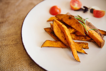 Close-up of a white ceramic plate with wedges of roasted organic sweet potato with rosemary in a...