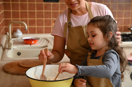 Caucasian cute little girl wearing a beige chefs apron, kneading a dough, standing near her mother...
