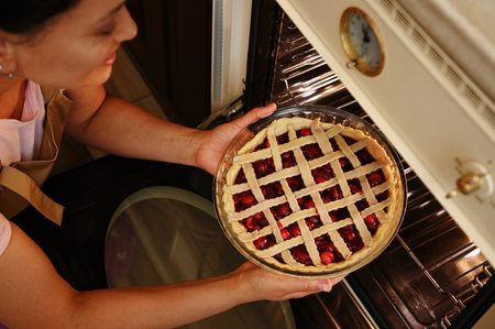 Close-up. View from above of a beautiful young woman putting baking dish with a cherry pie into oven...