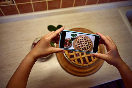 View from above of the hands of a female food blogger holding a smartphone and taking a picture of a classic American puff pastry cherry pie. Mobile phone in live view mode. Baking pastry itemsの写真素材