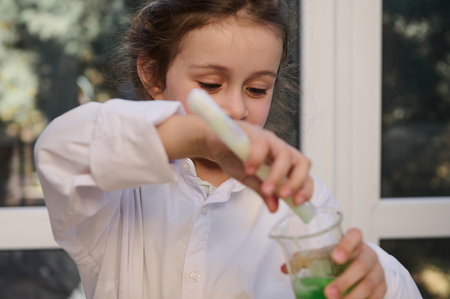 Focus on a mischievous little girl, a smart preschool child, in a white lab coat, conducting a...