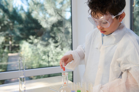 Smart school child, teenage boy in safety eyewear and lab coat, focused on doing scientific...