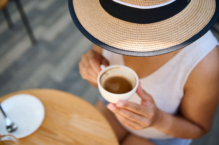 View from above of an unrecognizable young woman in straw hat and summer casual wear, holding a white cup of coffee americano, enjoying her coffee break outdoor, on a cozy summer terraceの写真素材