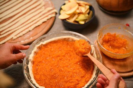 Close-up of a housewife holding a pumpkin puree spoon over a classic American Thanksgiving dinner pie. Fresh apple slices in ceramic bowl next to stripes of pastry for decorating a crispy lattice pieの写真素材