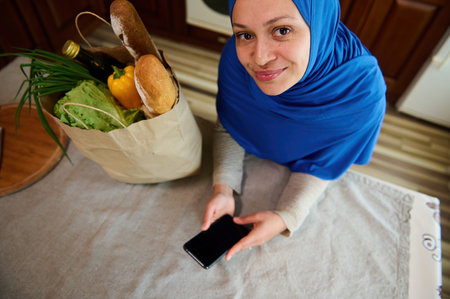 View from above of a pretty Middle-Eastern Muslim woman housewife in hijab, looks up at camera, checks expenses, using smartphone with copy advertising space for insert promotional text or mobile appの写真素材