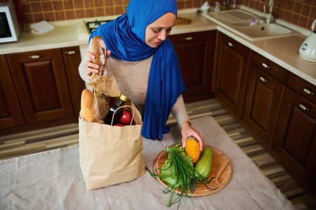 View from above of a Middle-Eastern Muslim woman in blue hijab and casual clothes, sorting fresh greens, organic vegetables while unpacking grocery shopping bag in the kitchen at homeの写真素材