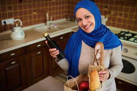 Pretty Middle-Eastern Muslim woman in blue hijab, sorting organic vegetables from a grocery shopping bag and putting them on the kitchen table, smiling a beautiful toothy smile looking at the cameraの写真素材