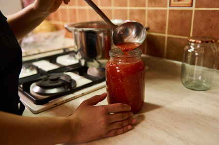 Close-up. Chef using a ladle, pours boiling juice of organic juicy tomatoes into a sterilized canning can. Preparing tomato sauce or passata. Preserving homegrown vegetables for winter. Canned foodの写真素材