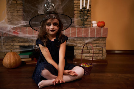 Cute little girl with Halloween themed make-up, dressed as a witch in a wizards hat, looks at camera, sitting against the backdrop of a fireplace covered with cobwebs, pumpkins and old mystical booksの写真素材