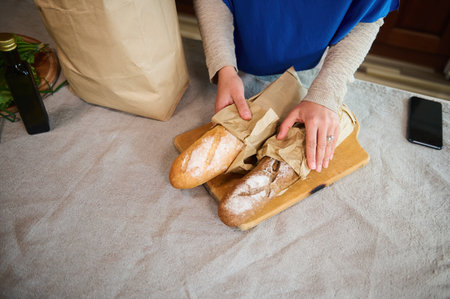 Top view of womans hands on baguettes of sourdough whole grain wheat bread while unpacking grocery bag in the home kitchen. Unrecognizable housewife unpacks shopping bag with groceries. Food deliveryの写真素材