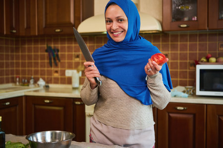 Charming Arabic Muslim woman housewife in blue hijab, smiling a toothy smile, looking at camera, standing with a kitchen knife and ripe fresh juicy tomato in her hands, against home kitchen backgroundの写真素材