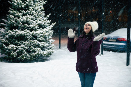 Joyful Latin American woman in warm clothes, enjoys the snowfall, rejoices in the first snow, standing on a snowy street illuminated by New Years garlands. Merry Christmas and happy winter holidaysの写真素材