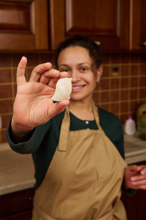 Selective focus. Carved bird from gingerbread dough in the hands of a blurred charming multi-ethnic woman housewife confectioner in beige apron, smiling a cheerful toothy smile looking at the cameraの写真素材