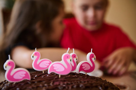 Selective focus on the pink flamingo candles, on the birthday cake with chocolate icing and nuts, on blurred background of charming diverse kids, having fun at birthday party. Celebration. Anniversaryの写真素材