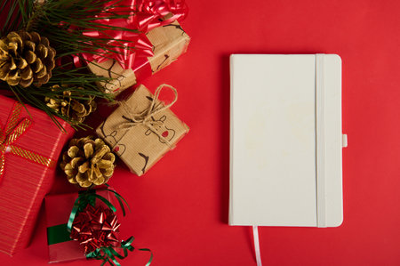 Flat lay. A white notepad or agenda book next to Christmas gift boxes, decorated with ribbons and bows and golden pine cones with fir-tree branch, isolated over red background. Copy ad space. Mockupの写真素材