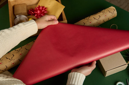 Top view womans hands rolling out red gift paper over green surface with laid out wrapping...