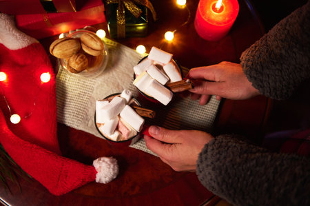 Overhead view of womans hands, putting hot cocoa drink with marshmallows on the green napkin on a wooden festive Christmas table, with delicious homemade cookies and garland lights, in candlelightの写真素材