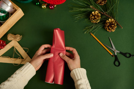 Close-up. Top view of a woman packaging a small present in red decorative paper over a green background with wrapping materials and golden pine cones as Christmas decor. Diy presents. Boxing Dayの写真素材