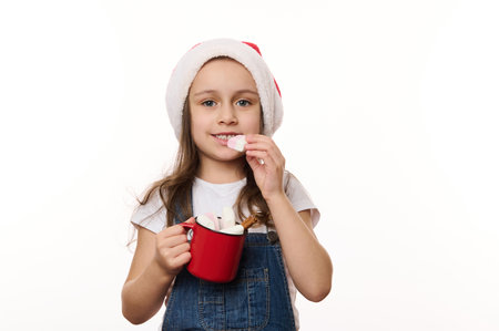 Isolated portrait on white background of lovely little child girl, in Santa hat, holding a red cup of hot chocolate and eating marshmallows. Magic atmosphere of upcoming Christmas, winter holidays.の写真素材