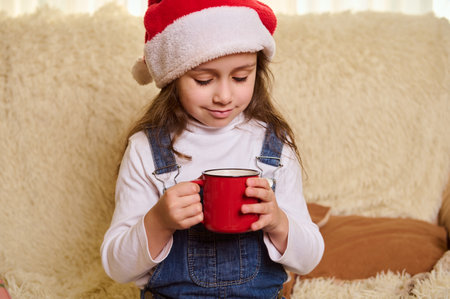 Adorable Caucasian child, adorable baby girl wearing Santa hat and denim overalls, holding a red cup of hot cocoa or chocolate drink with marshmallows, sitting on a comfortable sofa in the living roomの写真素材
