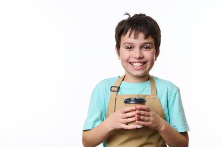 Multi-ethnic handsome preteen boy, wearing turquoise t-shirt and beige chefs apron holding an eco paper cup of takeaway coffee, smiling toothy smile looking at camera, isolated over white backgroundの写真素材