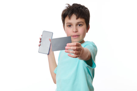 Focus on black credit card with free space for advertising text, in the hands of blurred school boy holding smartphone with blank screen, isolated on white backgroun. Internet banking. Online shoppingの写真素材