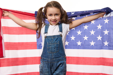 Happy Caucasian child, cute baby girl wearing blue denim overalls, smiling a beautiful toothy smile, looking at camera, holding American flag in her outstretched hands. 4th July - Independence Dayの写真素材