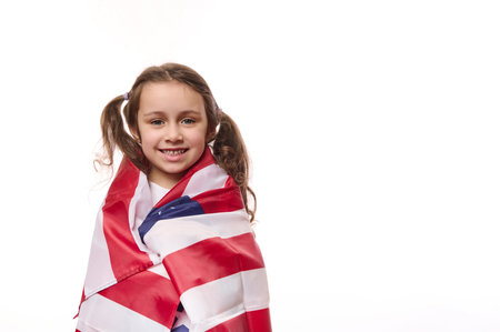 Lovely preschooler girl with two cute ponytails, wrapping in USA flag, celebrating the independence Day of United States of America on July 4 th. Concept of human rights and freedoms. White backgroundの写真素材