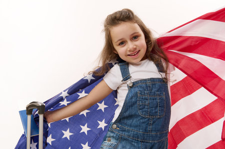 Happy baby girl wearing blue denim overalls with flag of USA, passport, boarding pass and suitcase, isolated over white background with free space for promotional text. Concept of travel, immigrationの写真素材