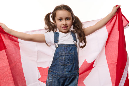 Happy little girl with two ponytails, wearing blue denim overalls, carries Canadian flag. Concept of Independence Day of Canada, the first July. Immigration. Emigration. Travel and tourism conceptの写真素材