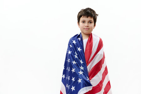 Confident American citizen, patriotic teenage boy wrapping in USA flag on the Independence Day of United States of America on July 4th, isolated over white background. Freedom and citizenship conceptの写真素材