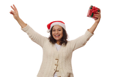 Beautiful overjoyed African American woman in Santa hat, holds a Christmas gift in her hands, cutely smiling looking at camera, isolated on white background. New Year celebration and Xmas conceptの写真素材