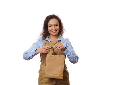 Pleasant multi-ethnic woman waitress in blue casual shirt and beige apron, packaging takeaway hot...