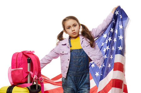 Little child girl pupil, smart primary school student in yellow pull, blue denim overalls and purple jacket, with backpack and suitcase, waving American flag, on white background. Education abroadの写真素材