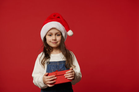 Lovely Caucasian little child girl in Santa hat, white pullover and blue denim overalls, smiling looking at camera, posing with cute gift box with surprise for Christmas, on red background. Ad spaceの写真素材
