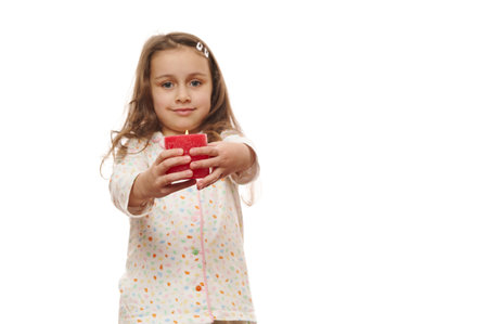 Focus on a red lit candle with fire flame, in hands of a blurred cute little child girl in comfortable soft and trendy pajamas, smiling cheerfully, looking at camera over white background. Copy spaceの写真素材